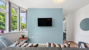 A modern living room with a large window, a flat-screen TV mounted on the wall, a grey sofa with striped cushions, and a small side table with a plant. - Modern 4 bed 3 bath apartment, free parking (Coventry)