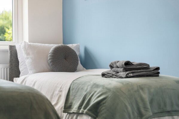 Un lit bien fait, avec des oreillers et une serviette pliée, une fenêtre donnant sur un paysage verdoyant et un mur bleu.
