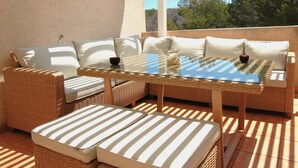 Un espace extérieur aménagé avec des meubles en osier, une table à plateau de verre et une vue sur les arbres et les collines.