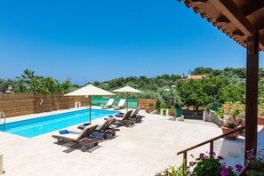 A swimming pool with lounge chairs and umbrellas, surrounded by greenery and a wooden fence. - Villa Aspa in Prines (Rethymno)