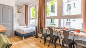 A room with a bed, a desk with chairs, a window with curtains, and a view of buildings outside. - WS Jardin du Luxembourg - Boissonade (Paris)