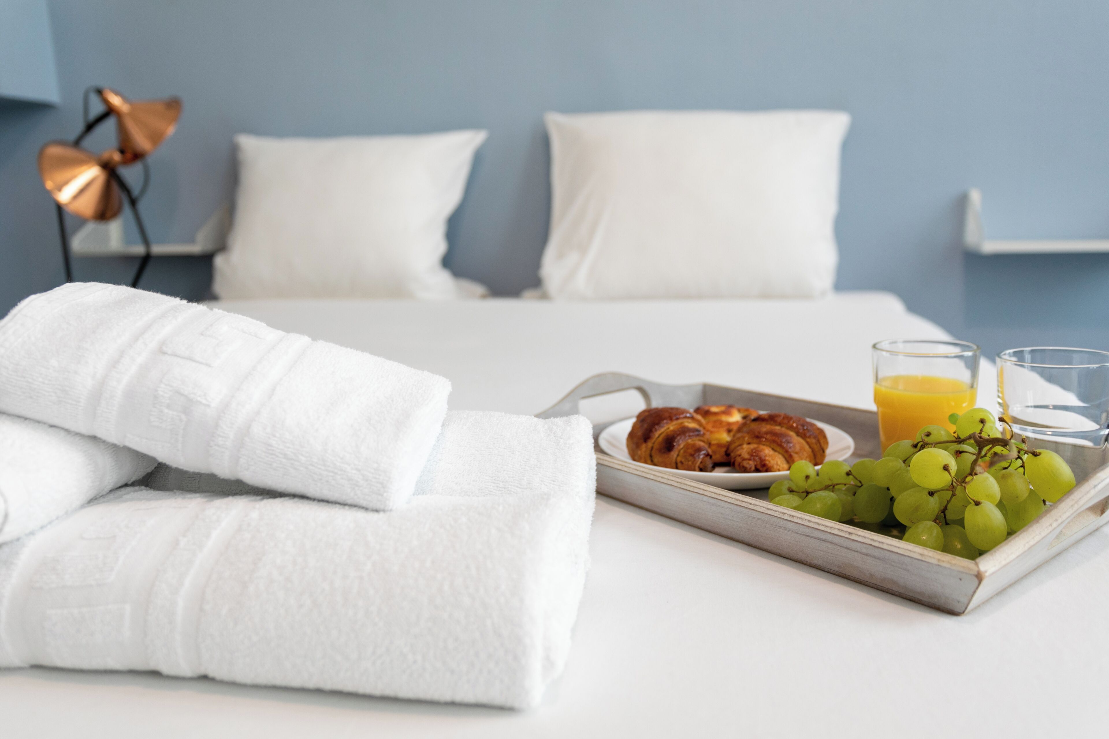 Una cama bien hecha con sábanas blancas, una bandeja con cruasanes, uvas y jugo de naranja, y una lámpara de noche.