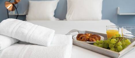 Una cama bien hecha con sábanas blancas, una bandeja con cruasanes, uvas y jugo de naranja, y una lámpara de noche.
