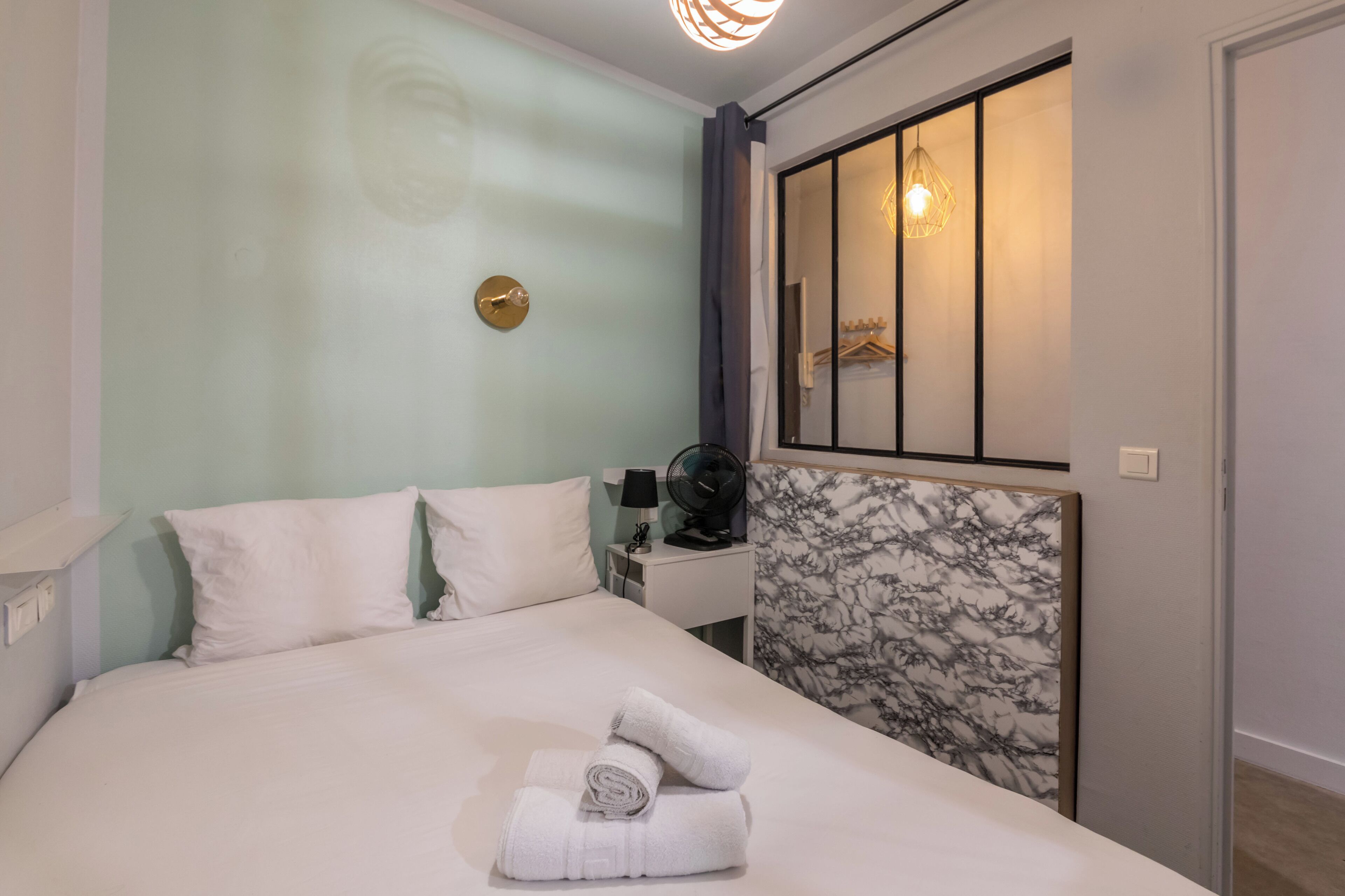Una habitación de hotel con cama, mesita de noche, ventilador, espejo y lámpara fijada a la pared.