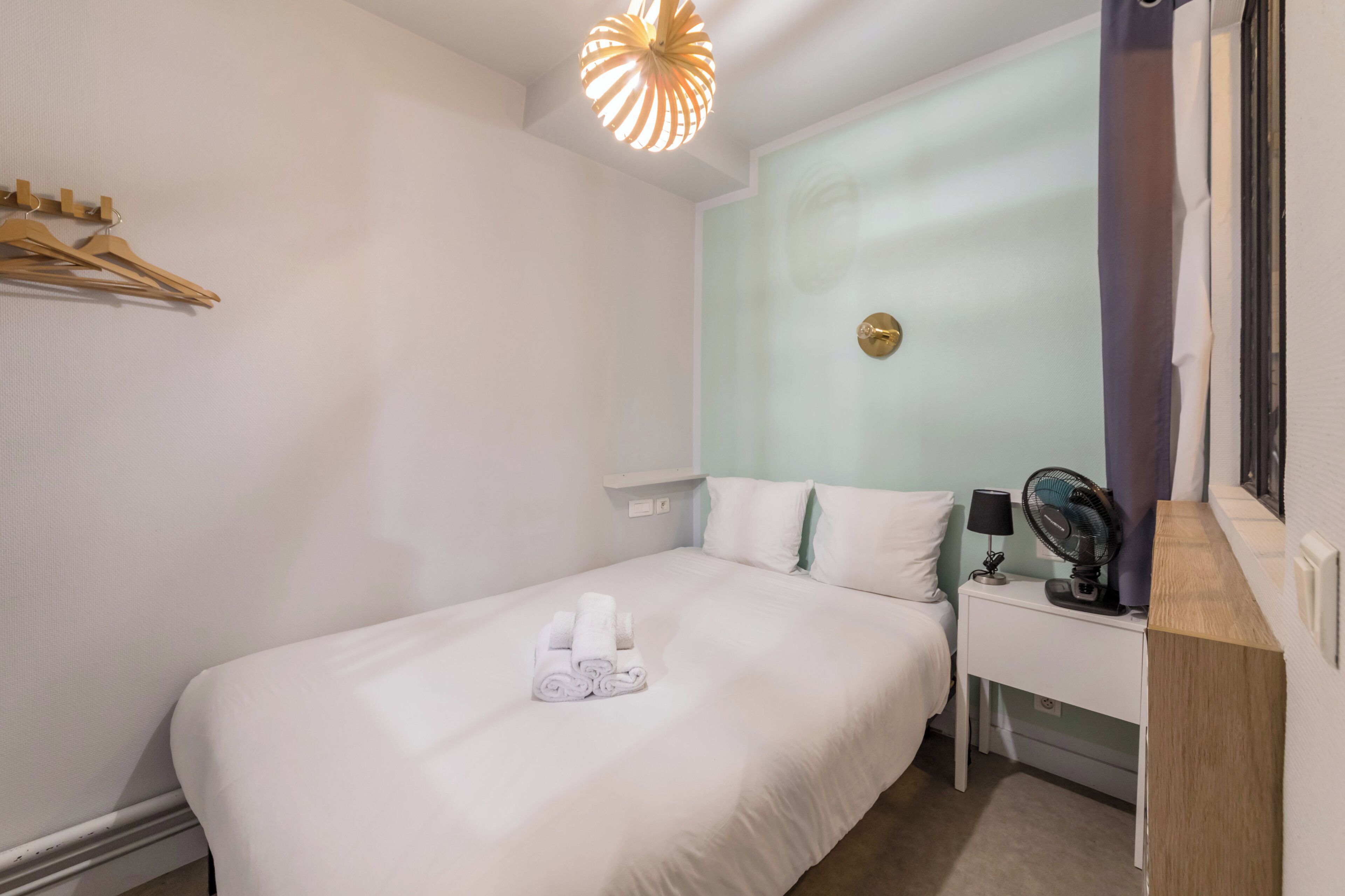 Una cama bien hecha con sábanas blancas y toallas enrolladas, una mesita de noche con lámpara y ventilador, y una lámpara fijada a la pared.