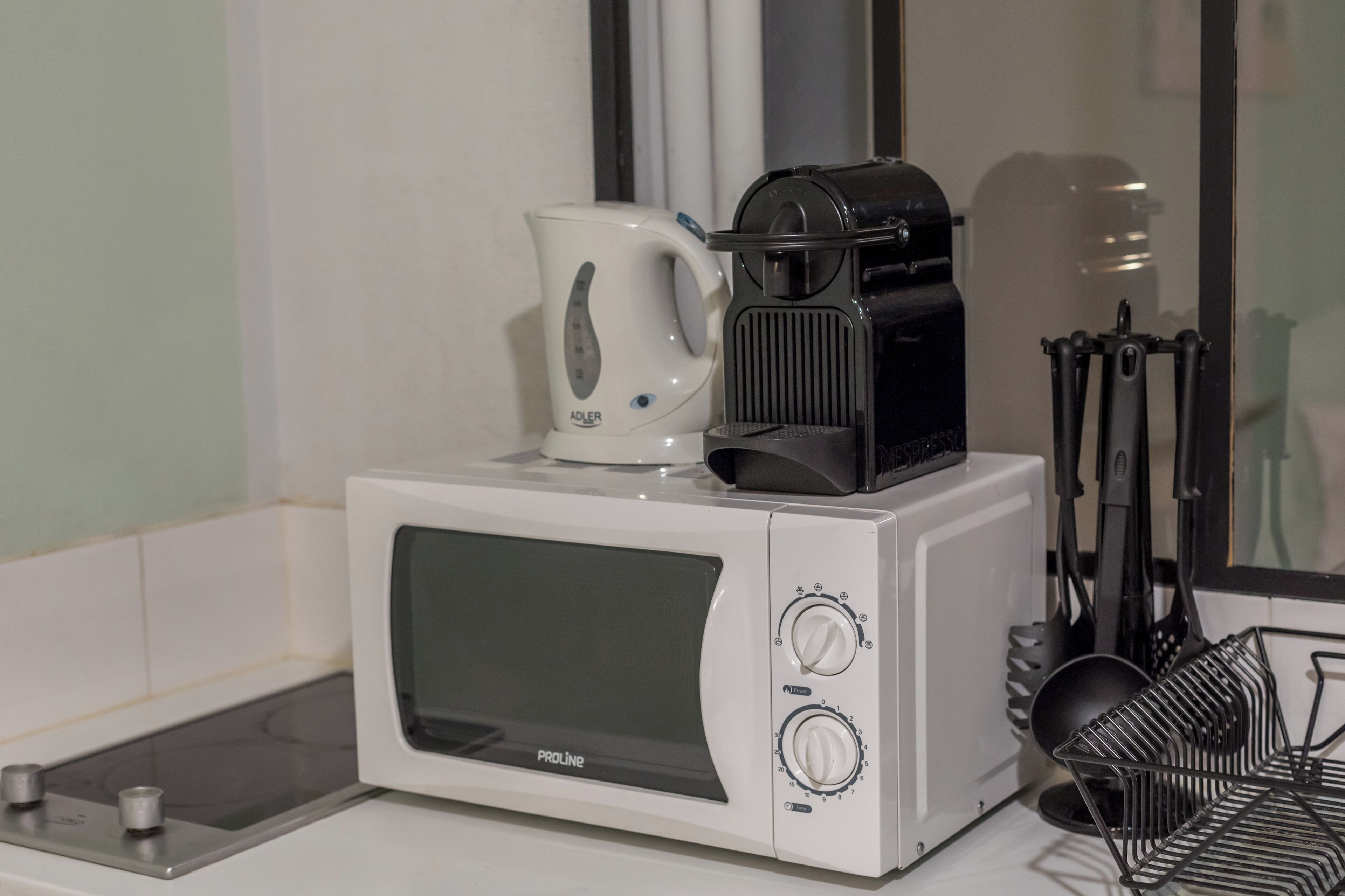 A compact kitchen with a white microwave, a coffee maker, and a black coffee pot on top. There are kitchen utensils hanging on the wall and a black metal rack with dishes.