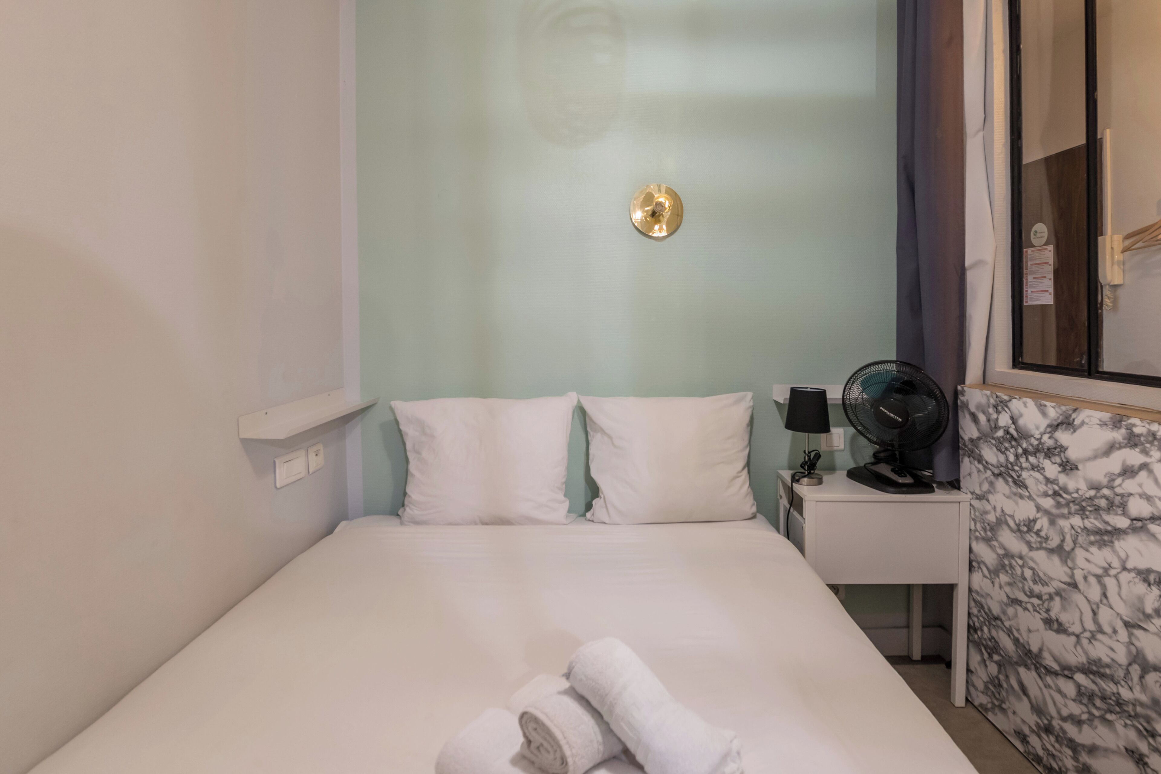 Habitación de hotel con cama, mesita de noche con lámpara y ventilador, espejo y pared con diseño de mármol.