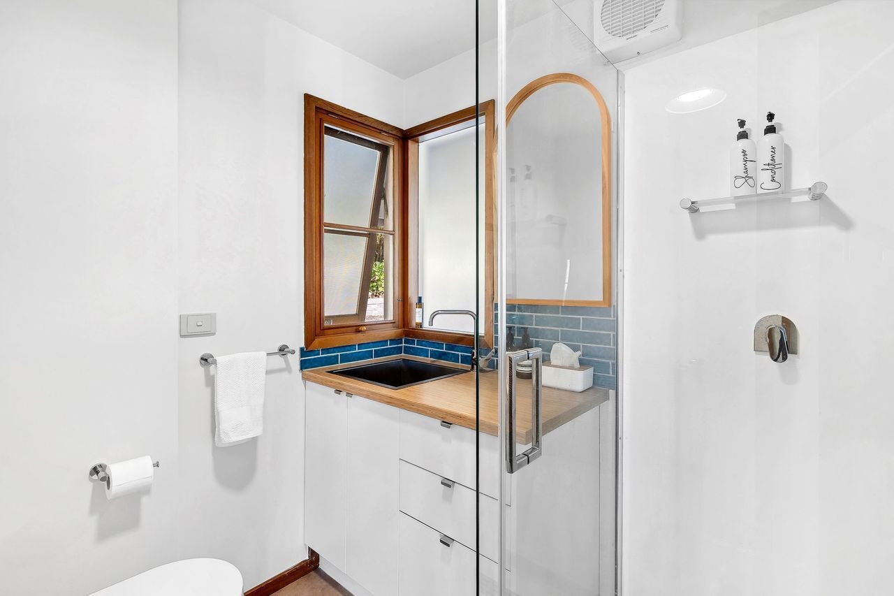 Un baño moderno con cabina de ducha de vidrio, ventana con marco de madera, lavamanos blanco y un salpicadero azul, y un perchero.