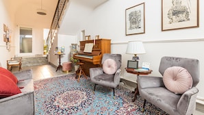 A spacious living room with a grand piano, two armchairs, a patterned rug, and a staircase leading to an upper floor.