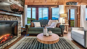 Ein gemütliches Wohnzimmer mit Kamin aus Stein, einem grünen Sofa und Blick auf Berge durch große Fenster.