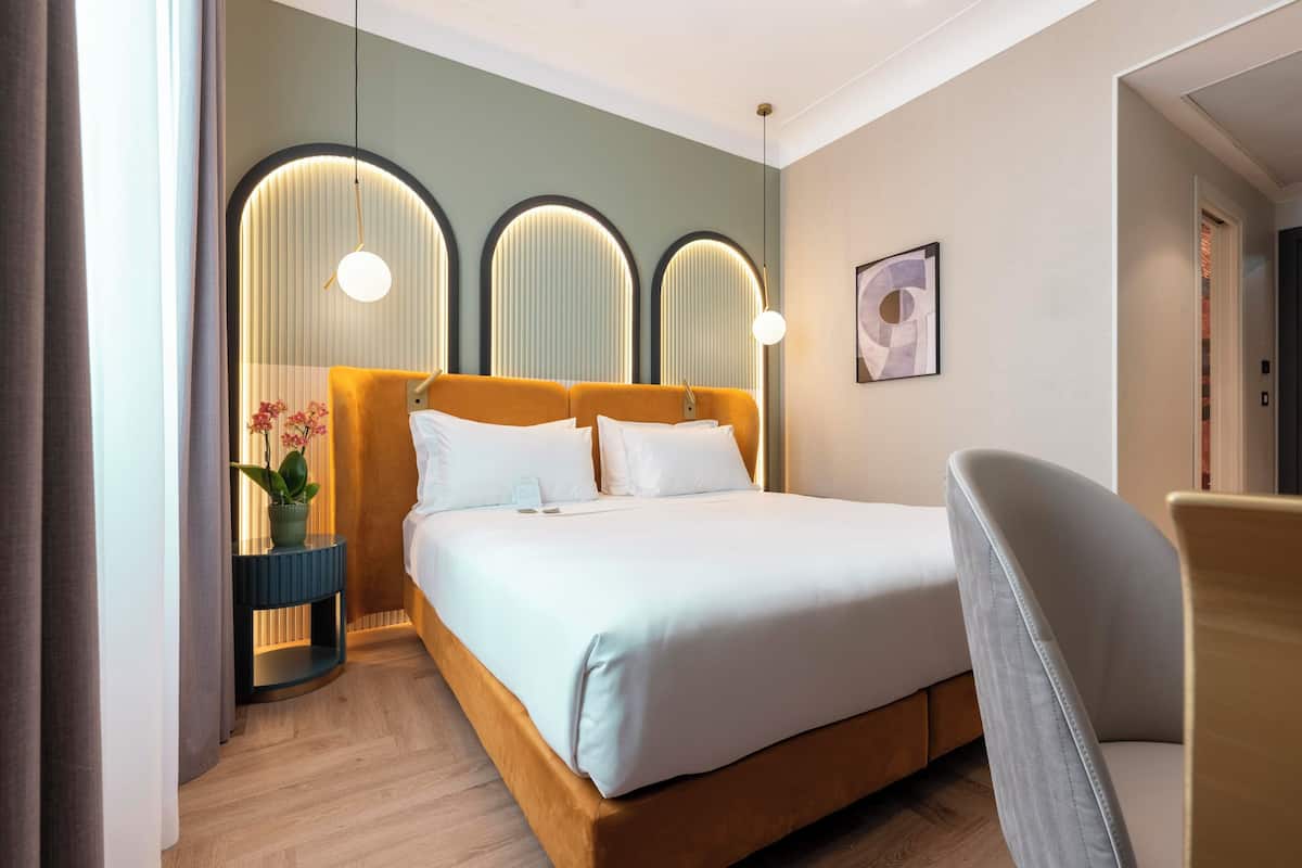 A modern hotel room with a large bed, a bedside table with a vase of flowers, and a chair.