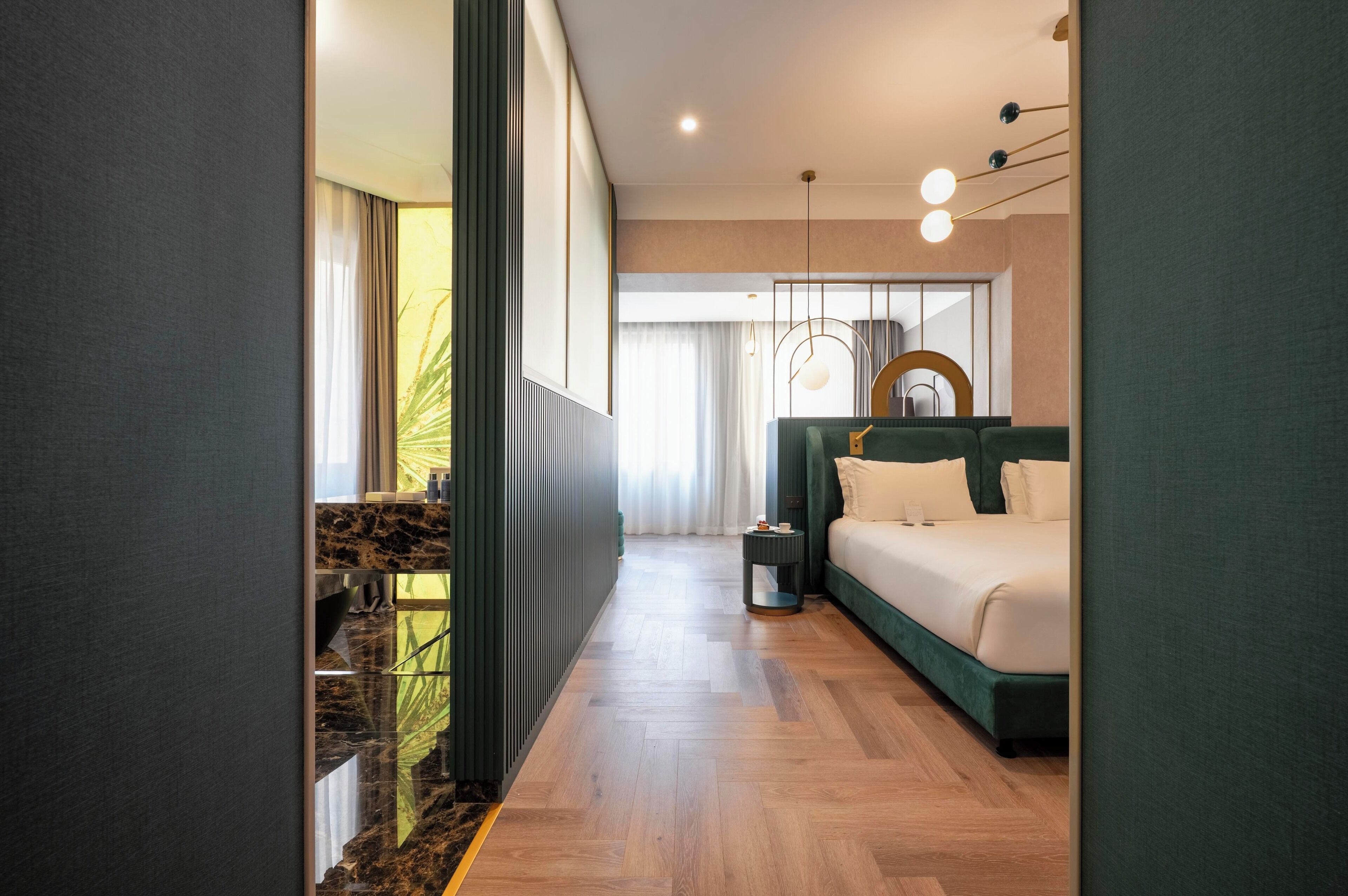 A modern hotel room with a wooden floor, a bed with green bedding, a nightstand, and a hanging light fixture.