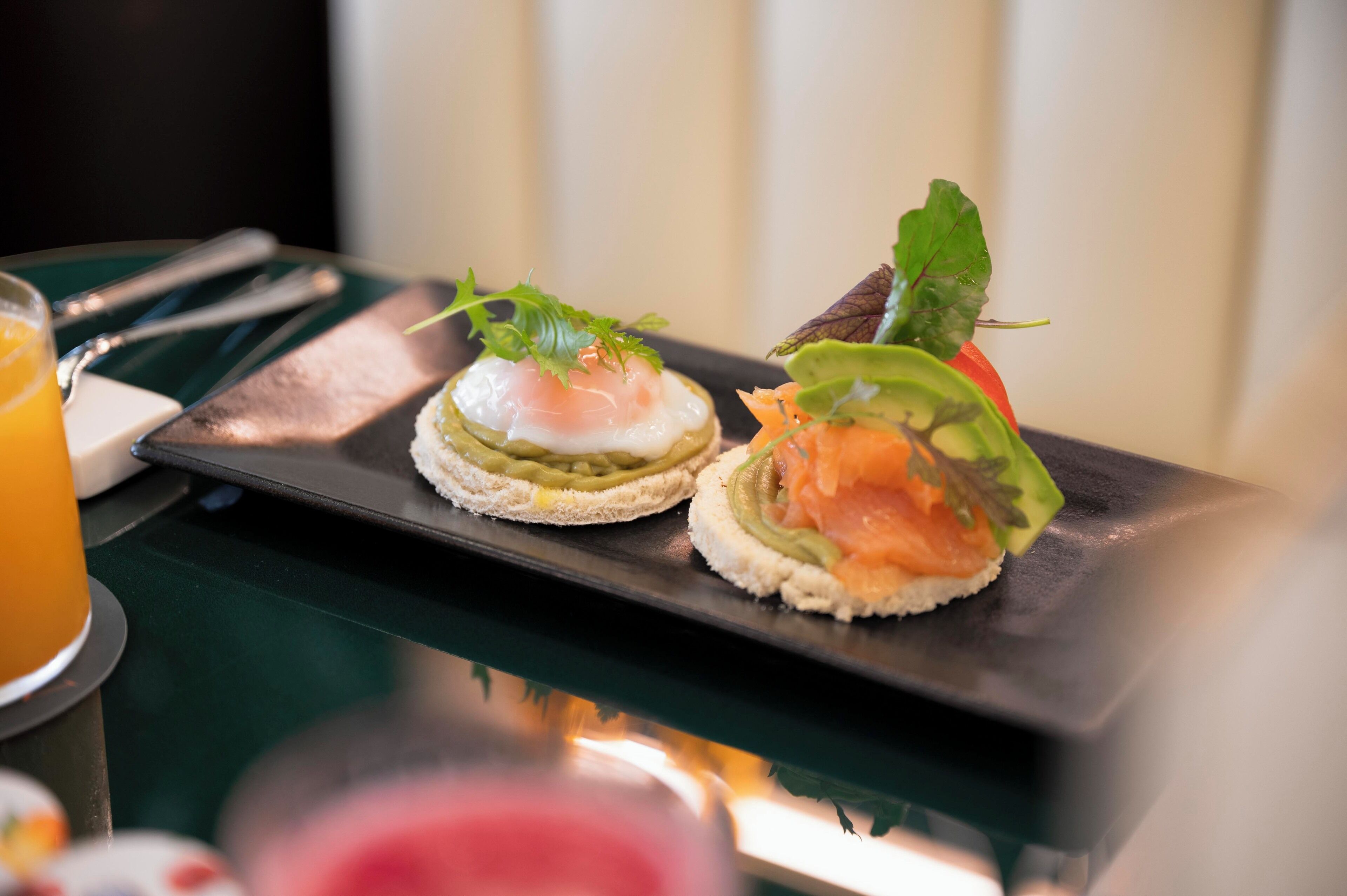Two open-faced sandwiches on a black rectangular plate, garnished with greens, on a table with a green surface.