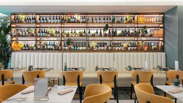 A restaurant with multiple tables and chairs, a wall of wine bottles, and a seating area with a cushioned bench.