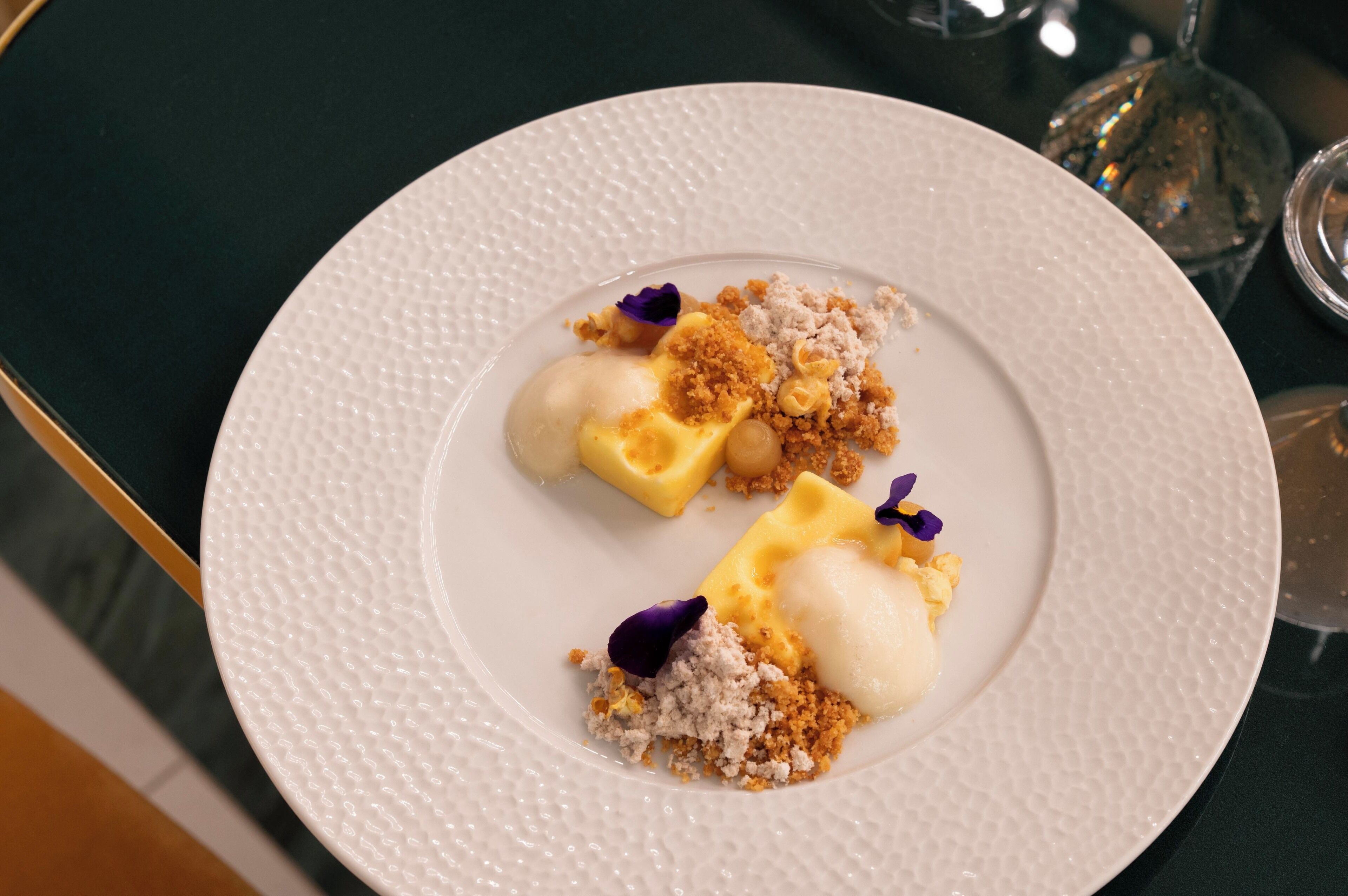 A dessert on a white textured plate with crumbled topping and purple garnish.