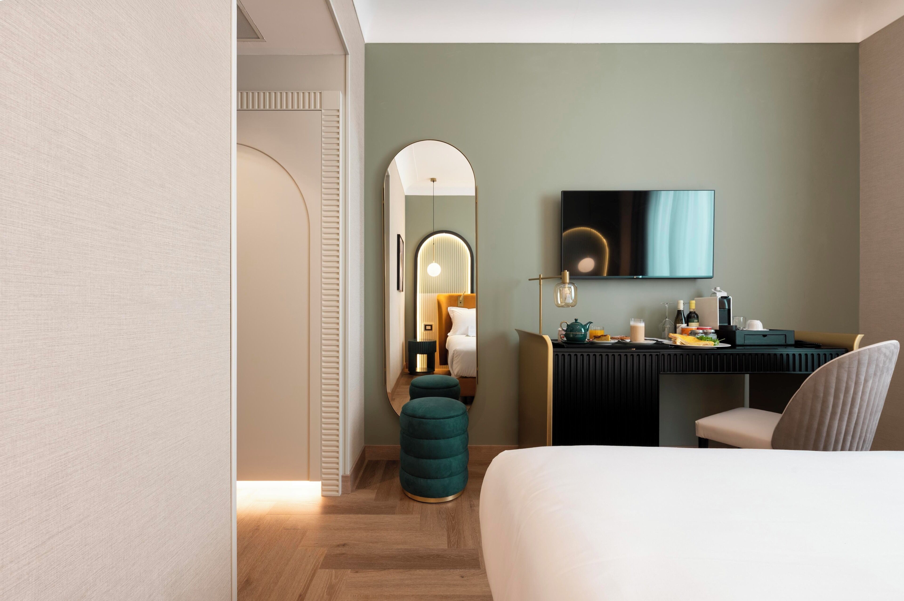 Quarto de hotel moderno com cama, escrivaninha com televisão de tela plana, espelho e um banquinho verde.