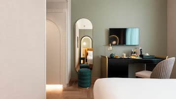Quarto de hotel moderno com cama, escrivaninha com televisão de tela plana, espelho e um banquinho verde.