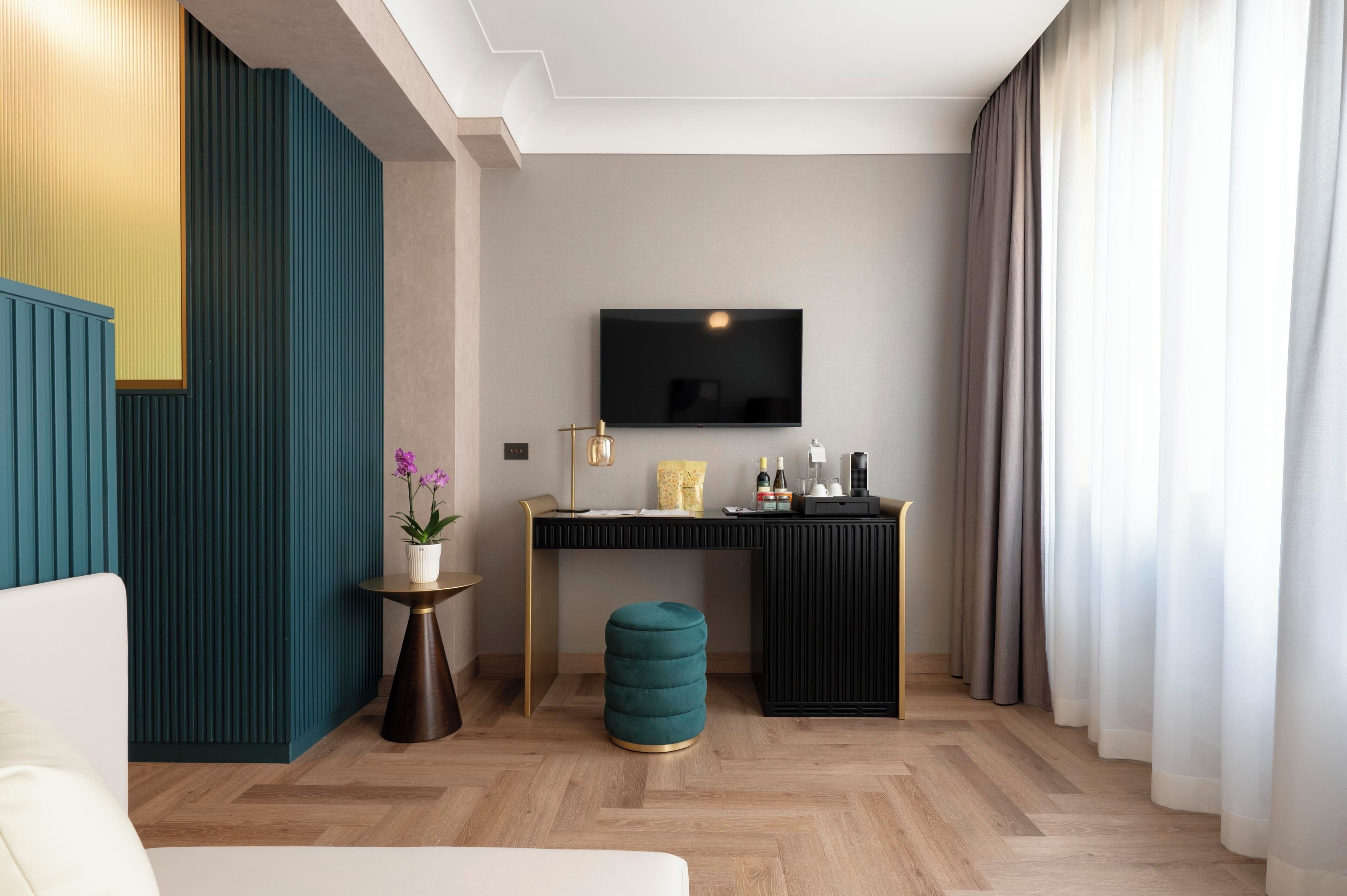 A modern hotel room with a flat-screen TV mounted on the wall, a wooden desk with a lamp, a small round ottoman, and a potted plant.