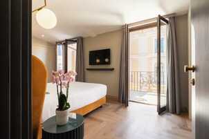 Una camera d'albergo con un letto, un tavolino con un vaso di fiori, una televisione a parete e un balcone con tende.