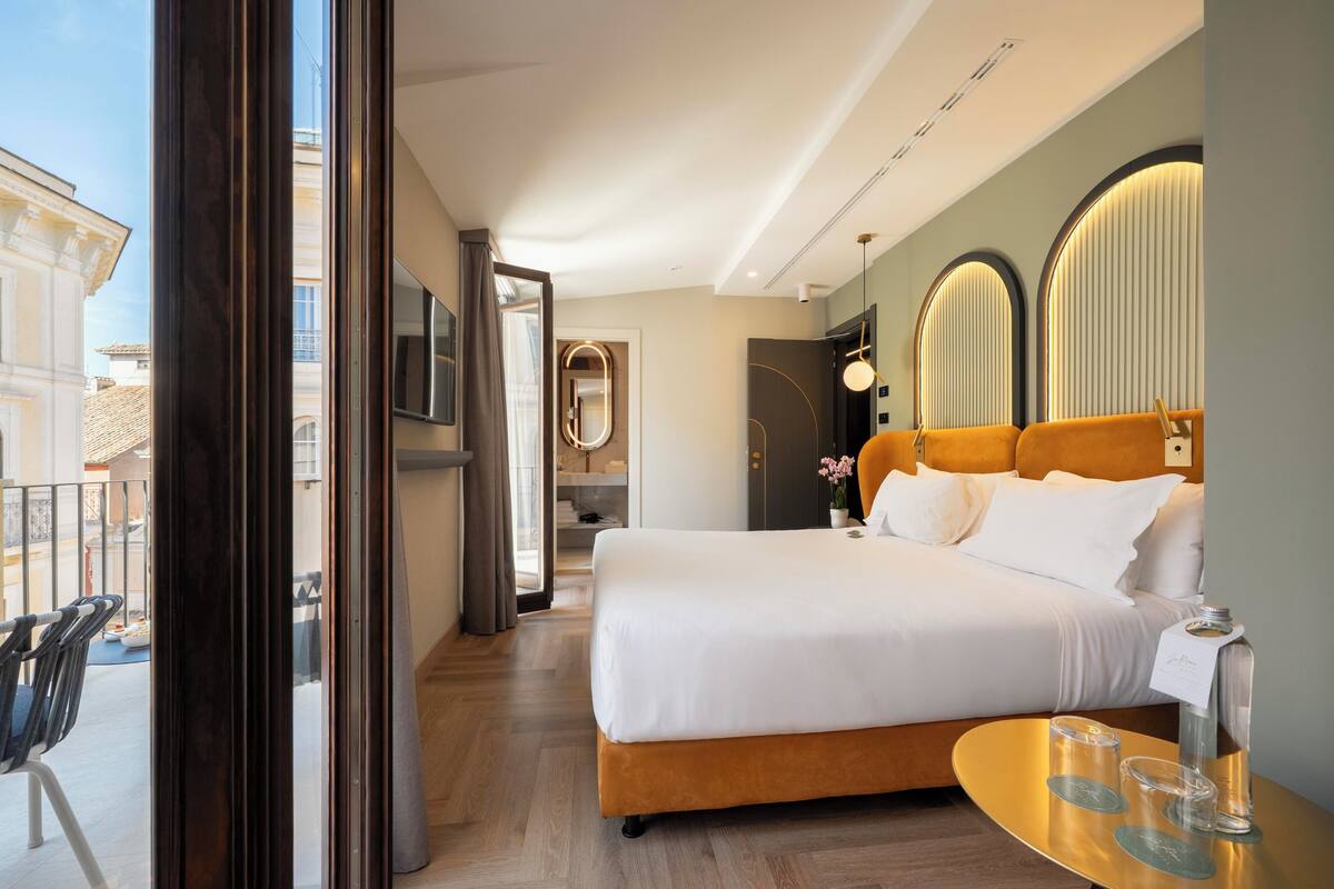 A hotel room with a large bed, a small table with glasses, a mirror, and a view of the outside balcony.