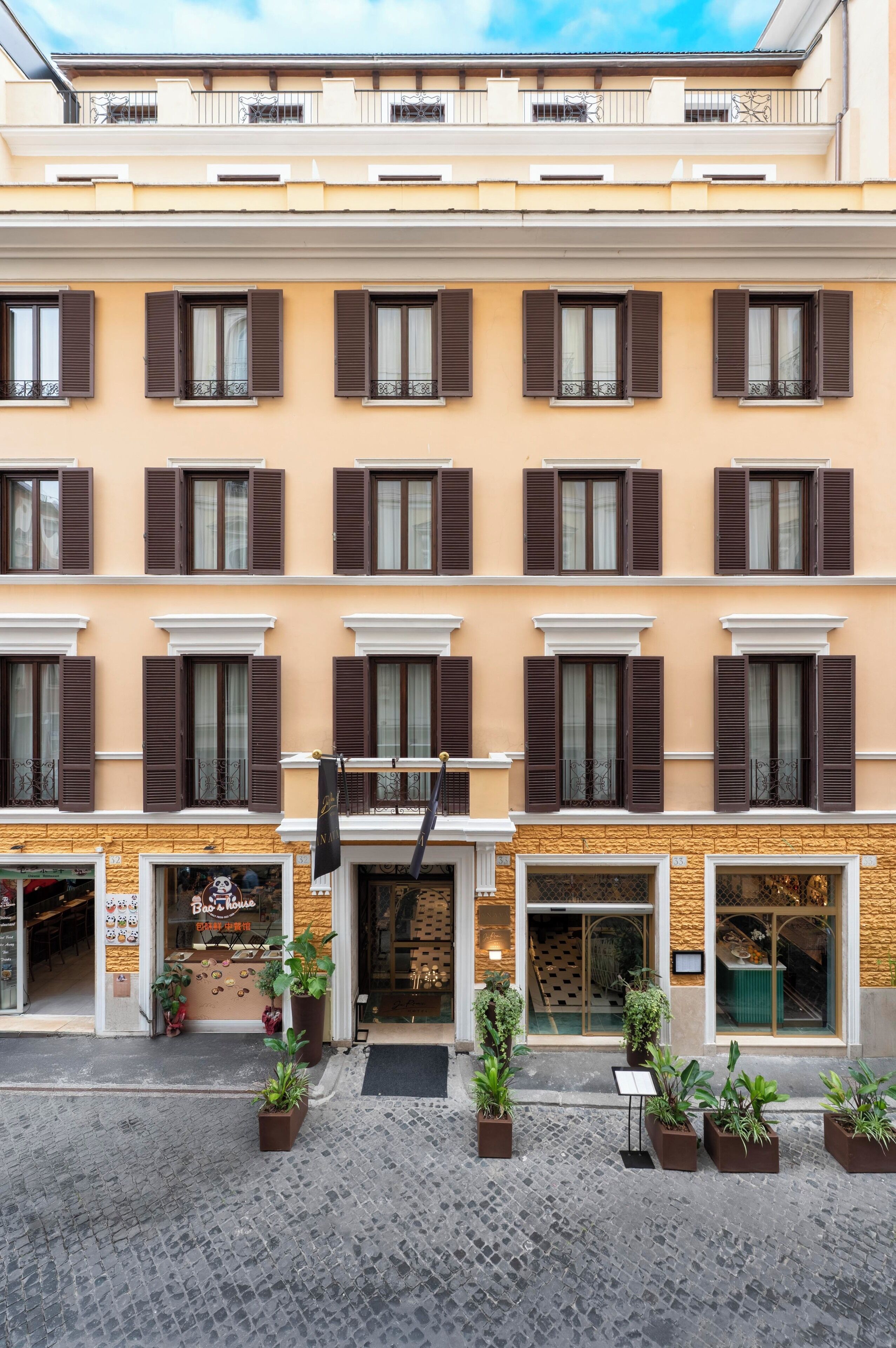 A multi-story building with a beige facade, brown shutters, and a cobblestone ground.