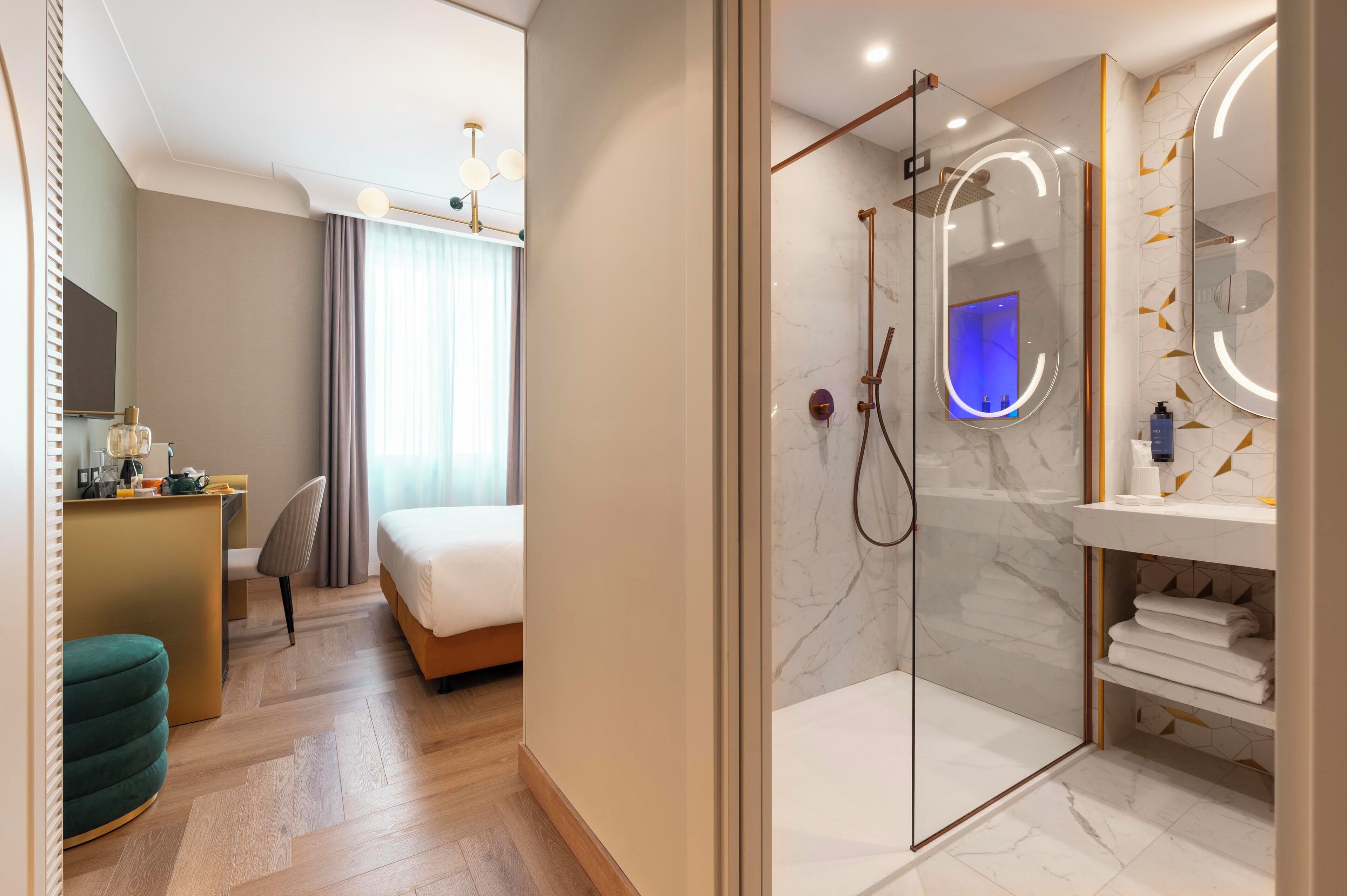 Una camera d'albergo moderna con un letto, una scrivania, un poggiapiedi verde, una doccia con porta in vetro e un bagno con parete in marmo e uno specchio.