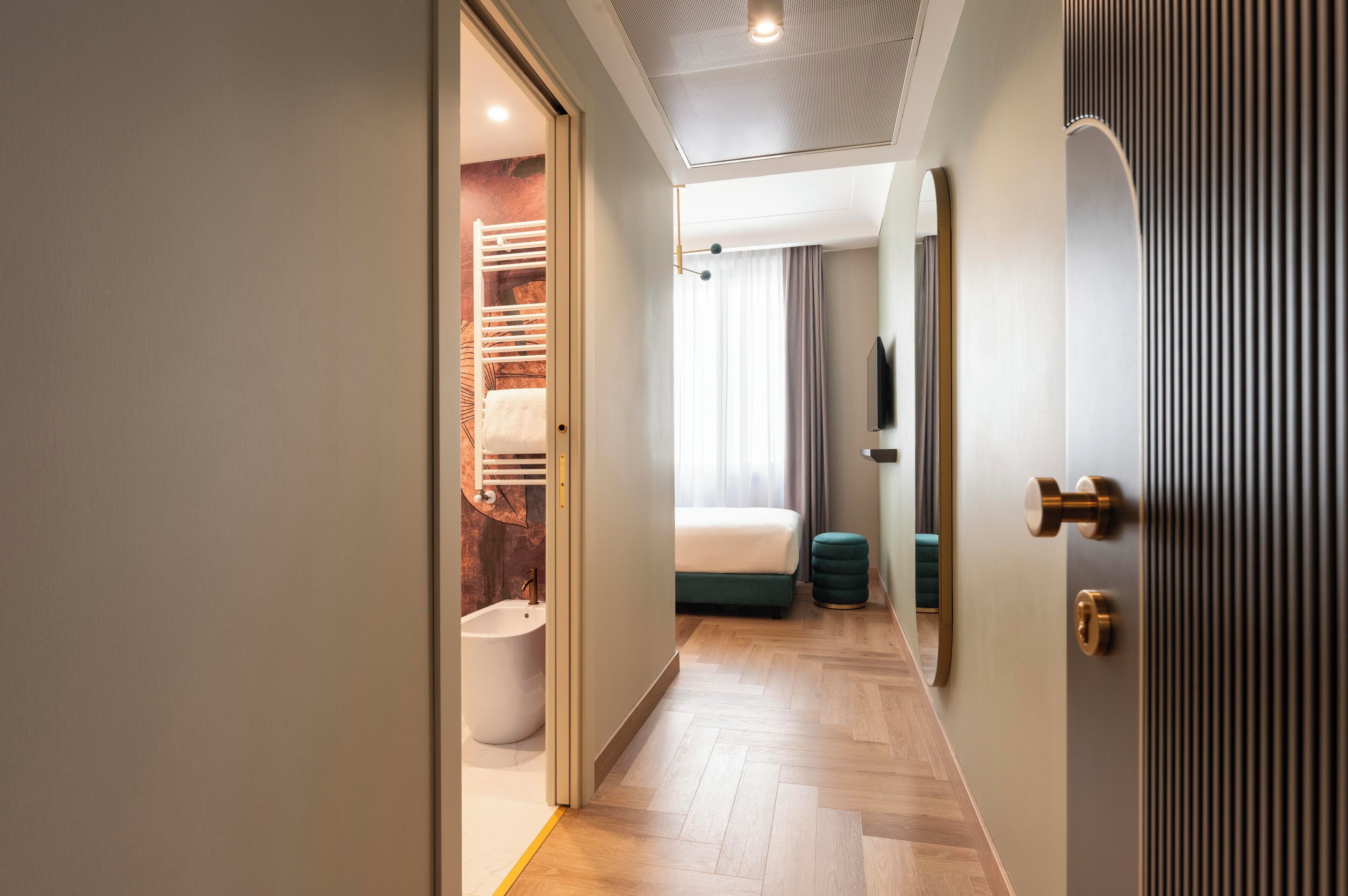 Una habitación de hotel moderna con piso de madera, una cama, un baño y un pasillo que conduce a una puerta.