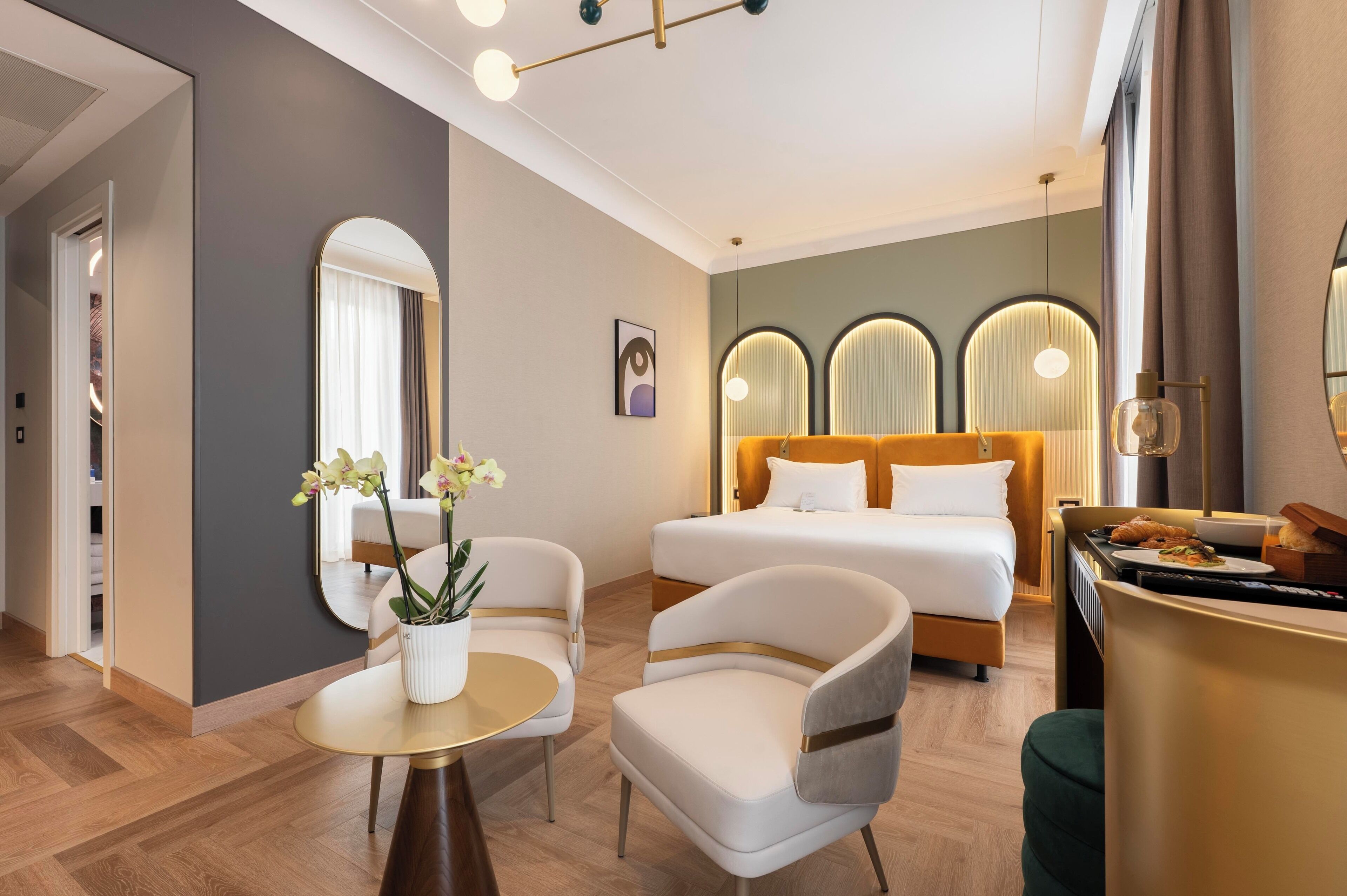 Una camera d'albergo con un letto, due poltrone, un tavolino, uno specchio e una finestra con tende.