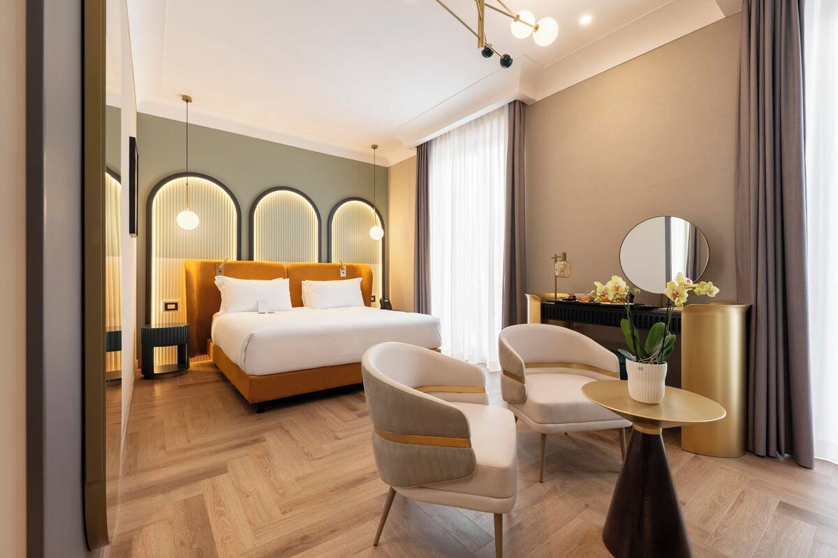 A modern hotel room with a bed, two chairs, a small table with a plant, a mirror, and a nightstand.