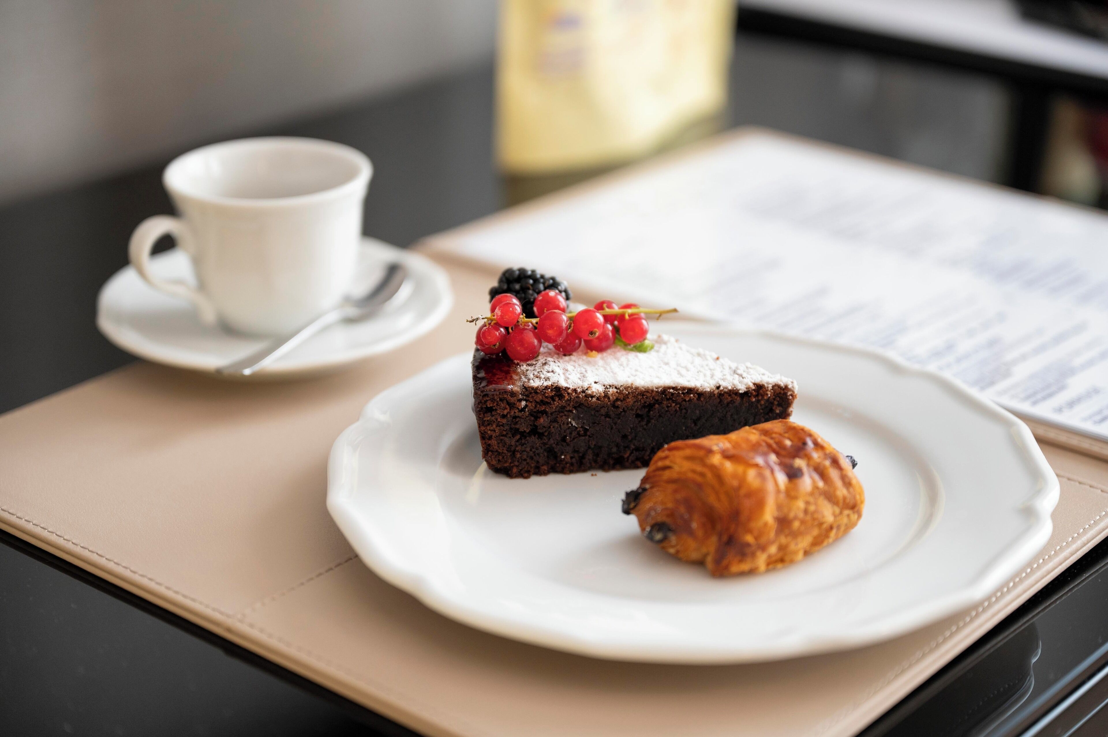 A dessert plate with a slice of cake topped with berries and powdered sugar, a croissant, and a white cup on a table with a menu.