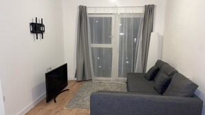 A modern living room with a grey sofa, a flat-screen TV, and a large window with sheer curtains. - New Cosy London Apartment With Garden View (London)
