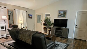 Una sala de estar con sofá de cuero negro, televisor de pantalla plana, chimenea y un ventanal con cortinas.