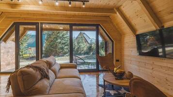 Ein gemütliches Zimmer mit großem Fenster, Holzbalkendecke, Ledersofa, Esstisch mit Stühlen und einem Fernseher an der Wand.