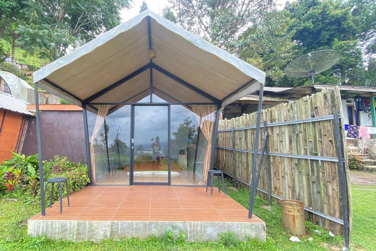 A covered outdoor seating area with a glass wall, surrounded by greenery and a wooden fence.