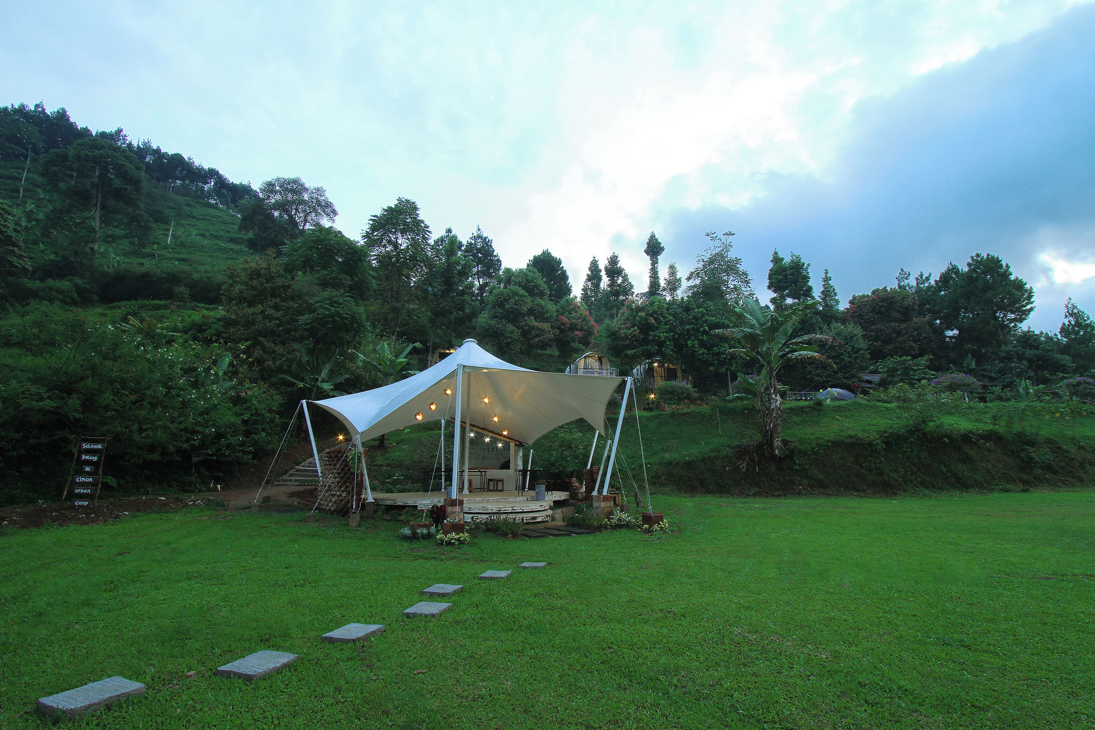 A tented outdoor area with a stone pathway leading to it, surrounded by lush greenery and hills.
