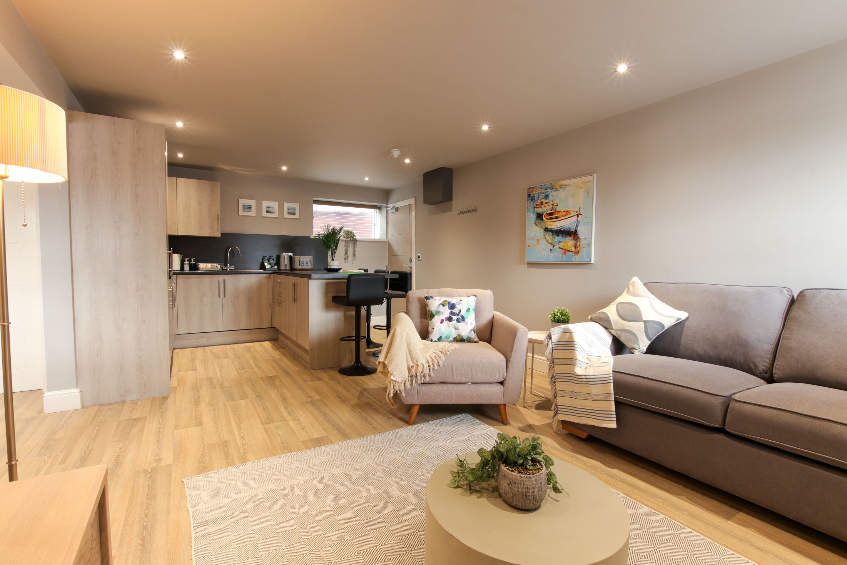 Un espace de vie moderne avec un canapé, un fauteuil et une table basse, une cuisine en arrière-plan et un tableau accroché au mur.