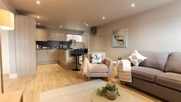 Un espace de vie moderne avec un canapé, un fauteuil et une table basse, une cuisine en arrière-plan et un tableau accroché au mur.