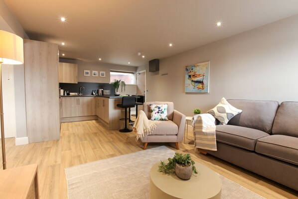 A modern living area with a sofa, armchair, and coffee table, a kitchen in the background, and a painting on the wall. - The Ironworks Aparthotel (Telford)