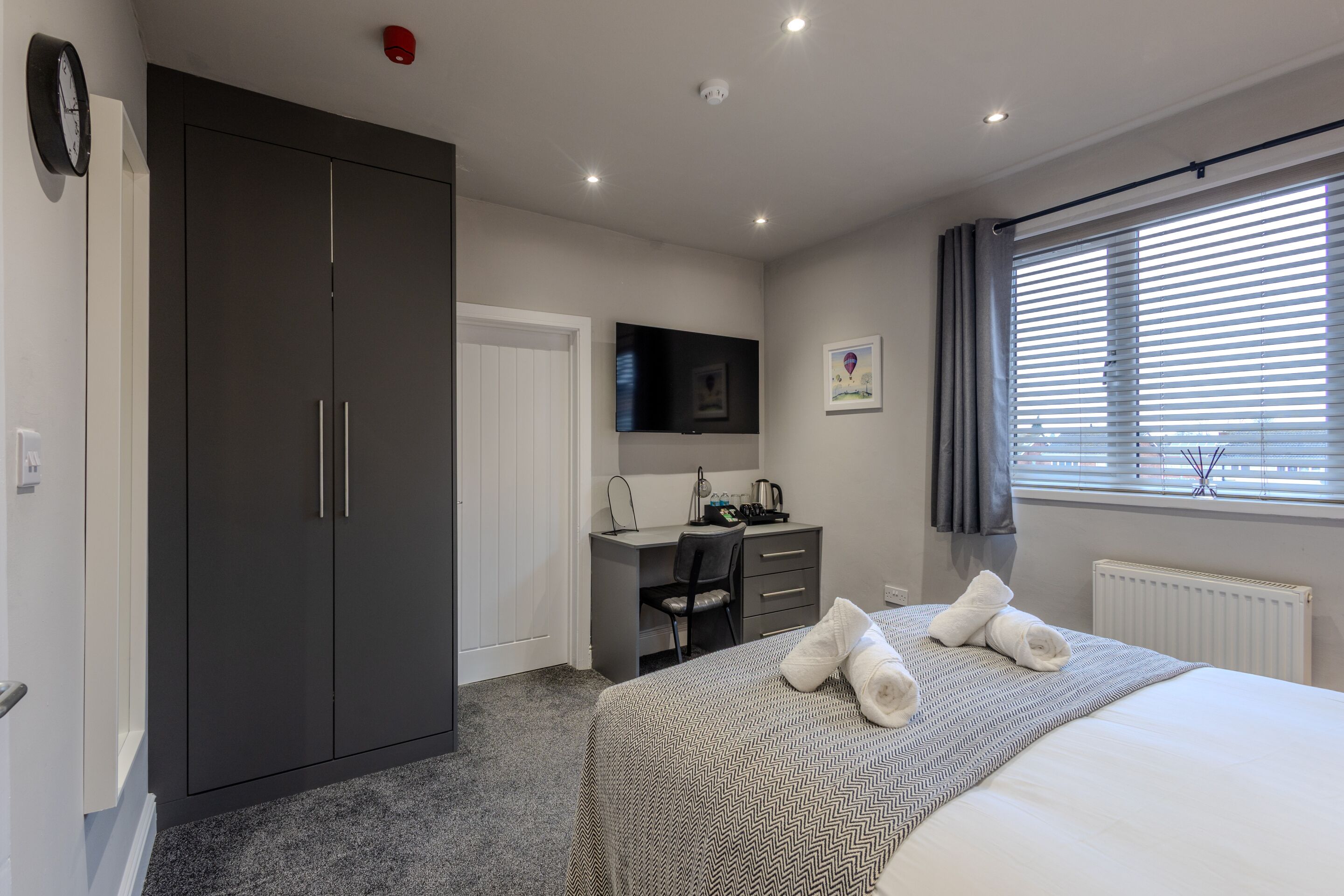 Une chambre à coucher comprenant un lit, un bureau, une chaise, une armoire, un téléviseur et une fenêtre avec des stores.