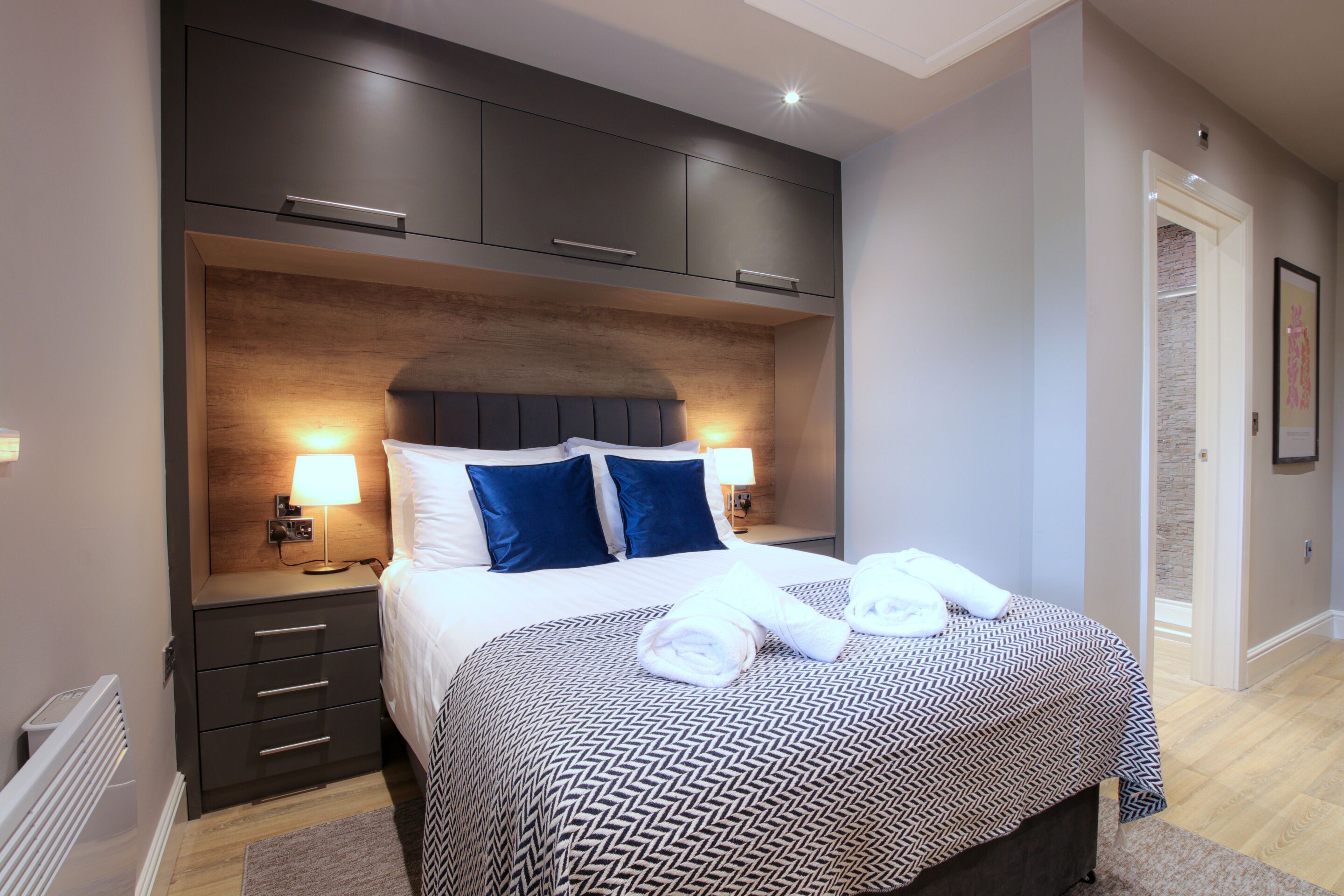 A modern hotel room with a large bed, dark wood headboard, and a patterned bedspread. There are two bedside lamps and a nightstand with a lamp.