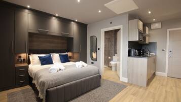 A modern bedroom with a large bed, dark cabinetry, and a small kitchenette.