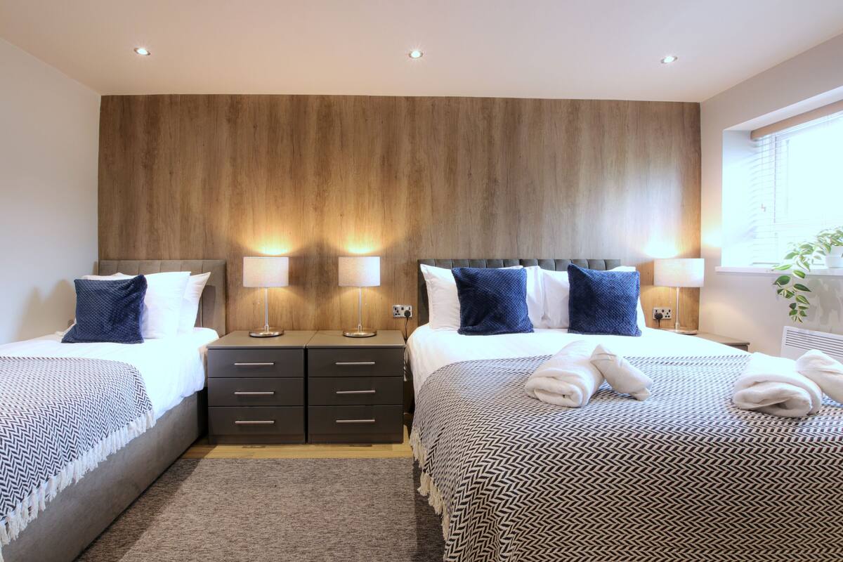 A modern hotel room with two beds, wooden headboard, bedside lamps, and a large window with blinds.