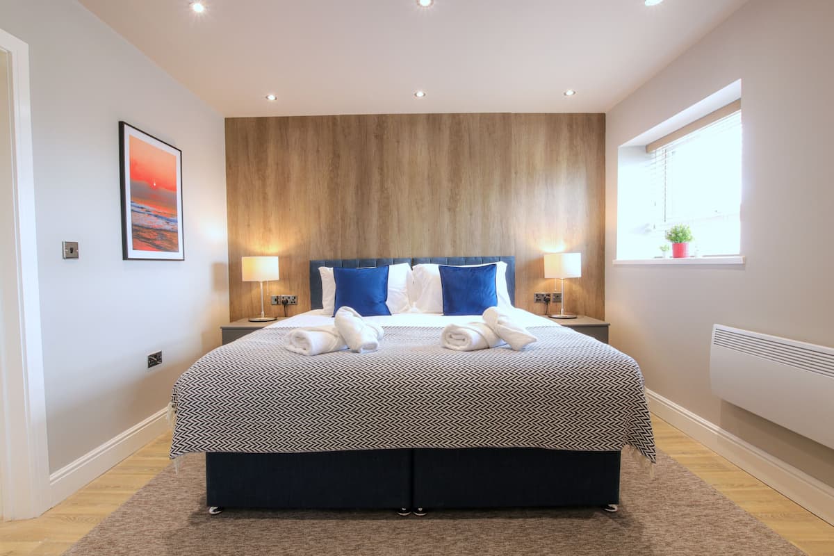 A modern bedroom with a large bed, two bedside lamps, a wooden headboard, and a window with a plant.