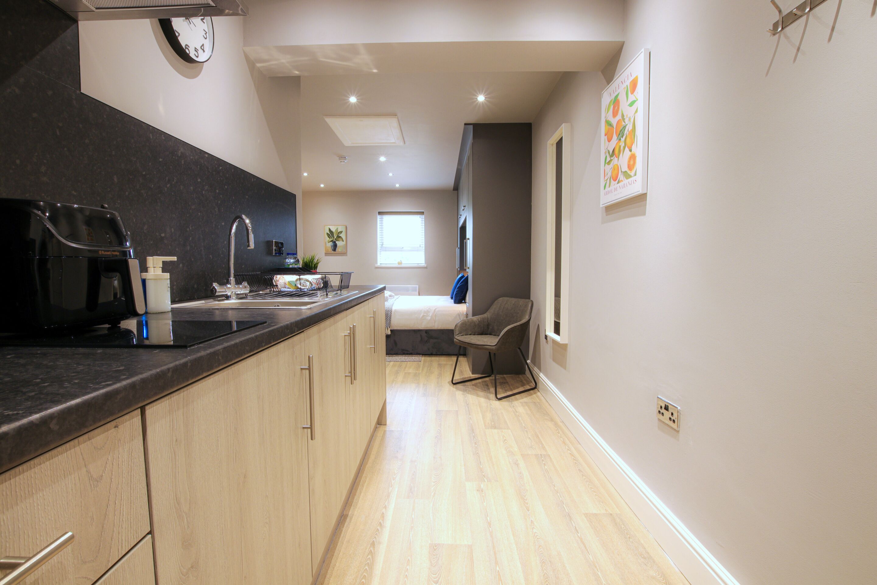 A modern kitchen with a black countertop, wooden cabinets, and a built-in microwave. There is a window with blinds and a chair in the adjacent room.