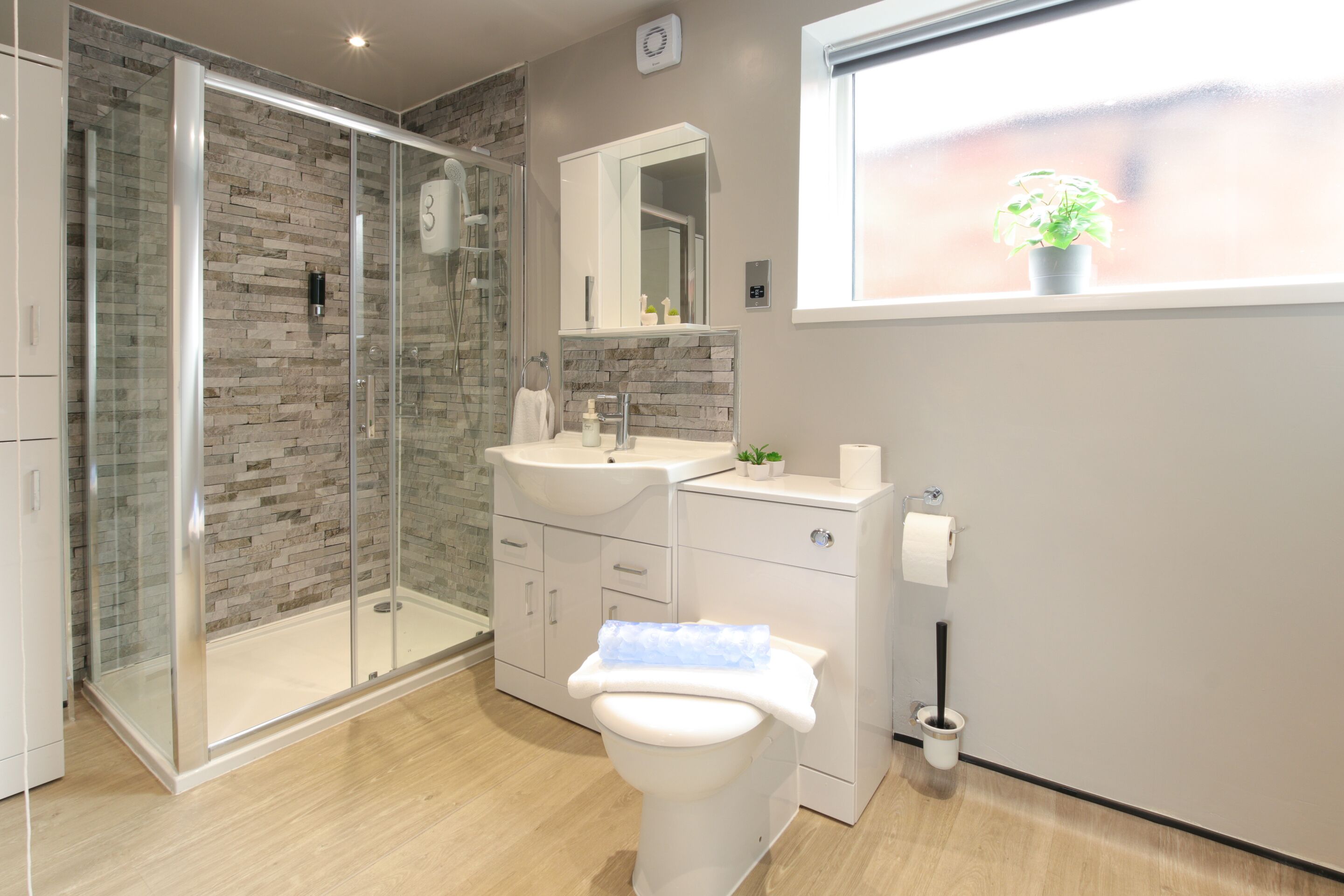 A modern bathroom with a glass-enclosed shower, a white toilet, a sink with a mirror above, and a window with a plant.