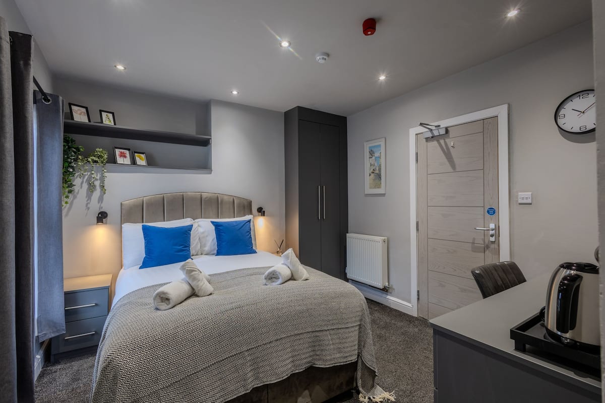 A modern hotel room with a bed, bedside table, and a desk with a kettle.