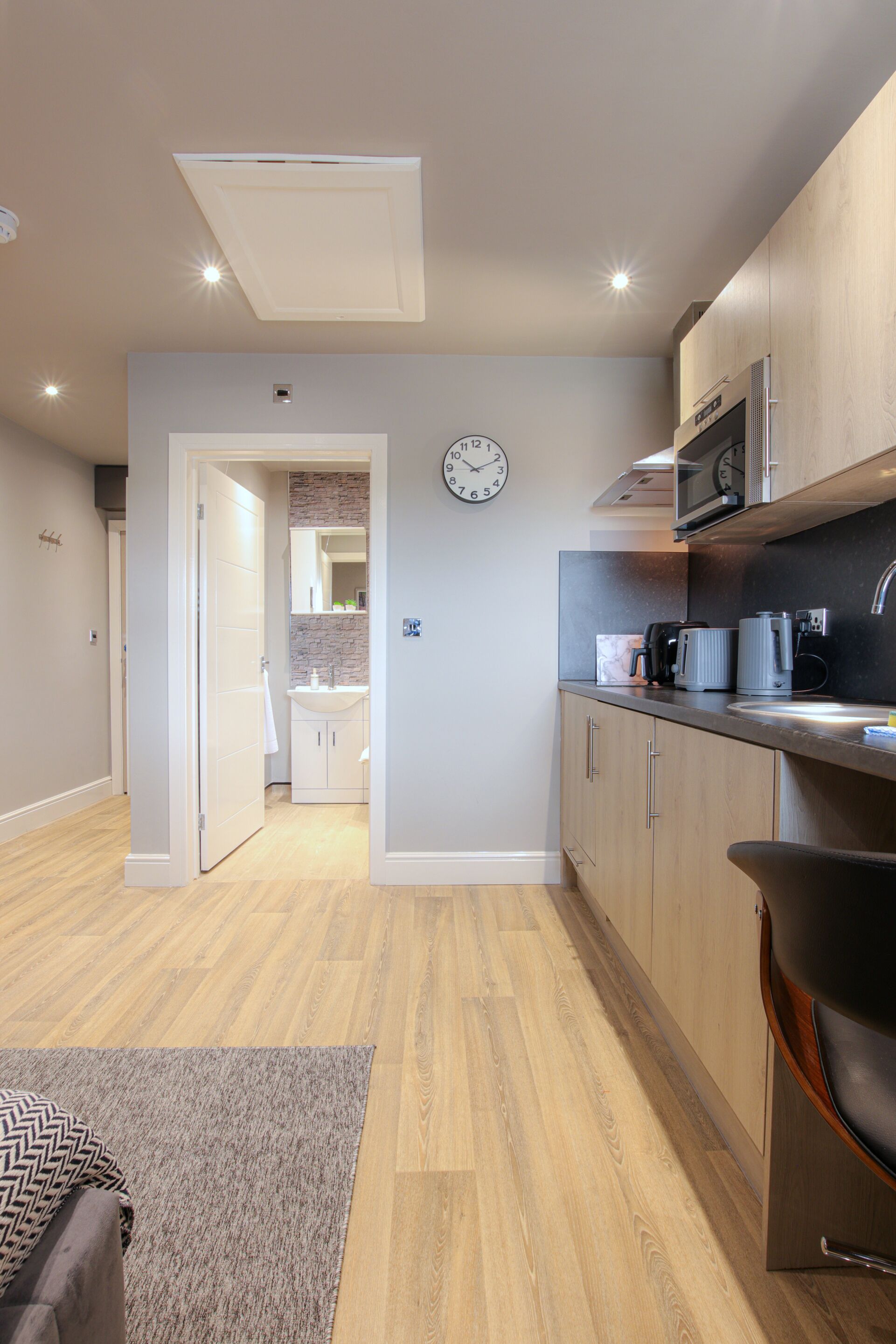 A modern kitchen with wooden cabinets, a built-in microwave, and a sink. There is a clock on the wall and a door leading to another room.