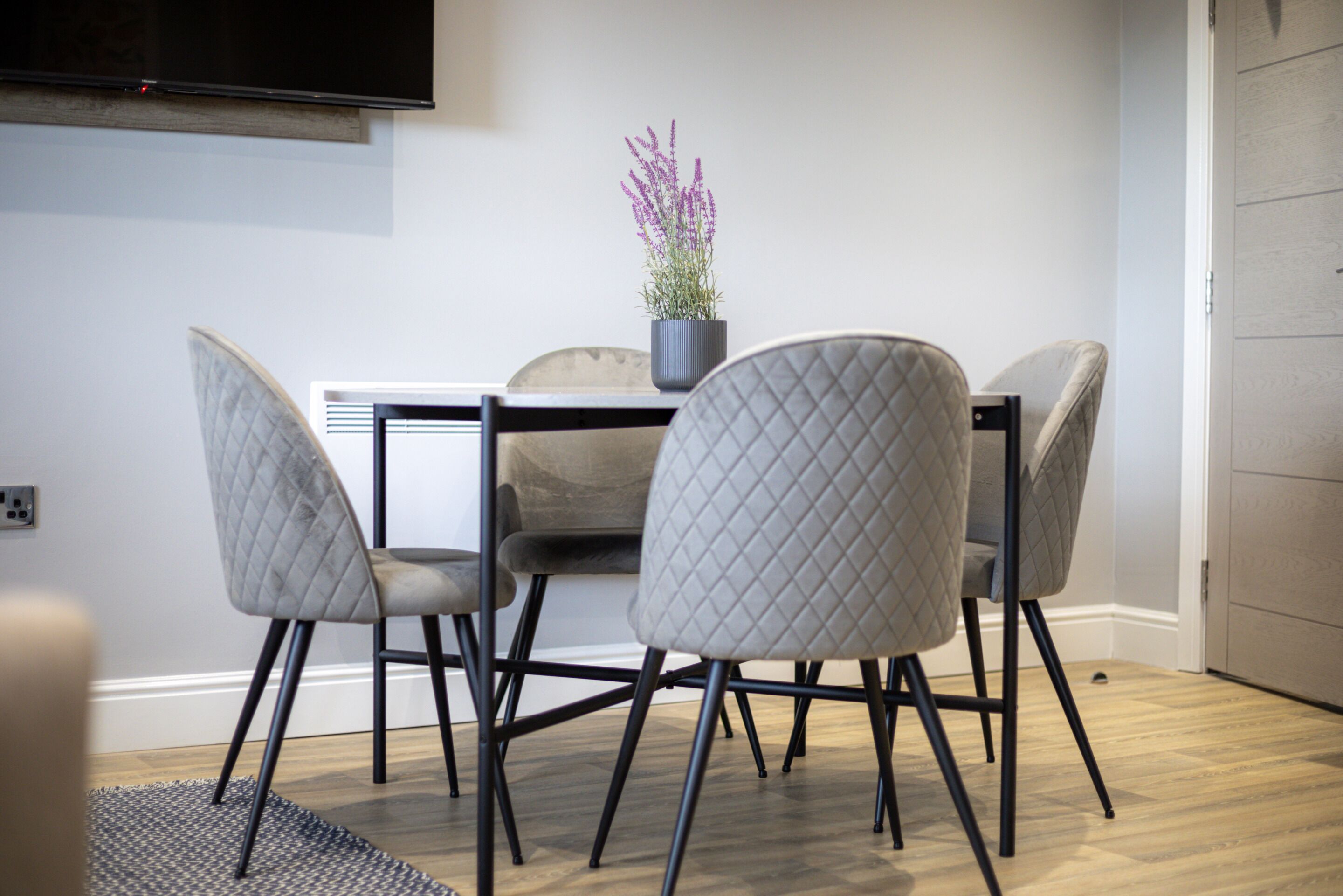 Uma área de refeições com mesa e quatro cadeiras, um vaso de planta e uma televisão de tela plana na parede.