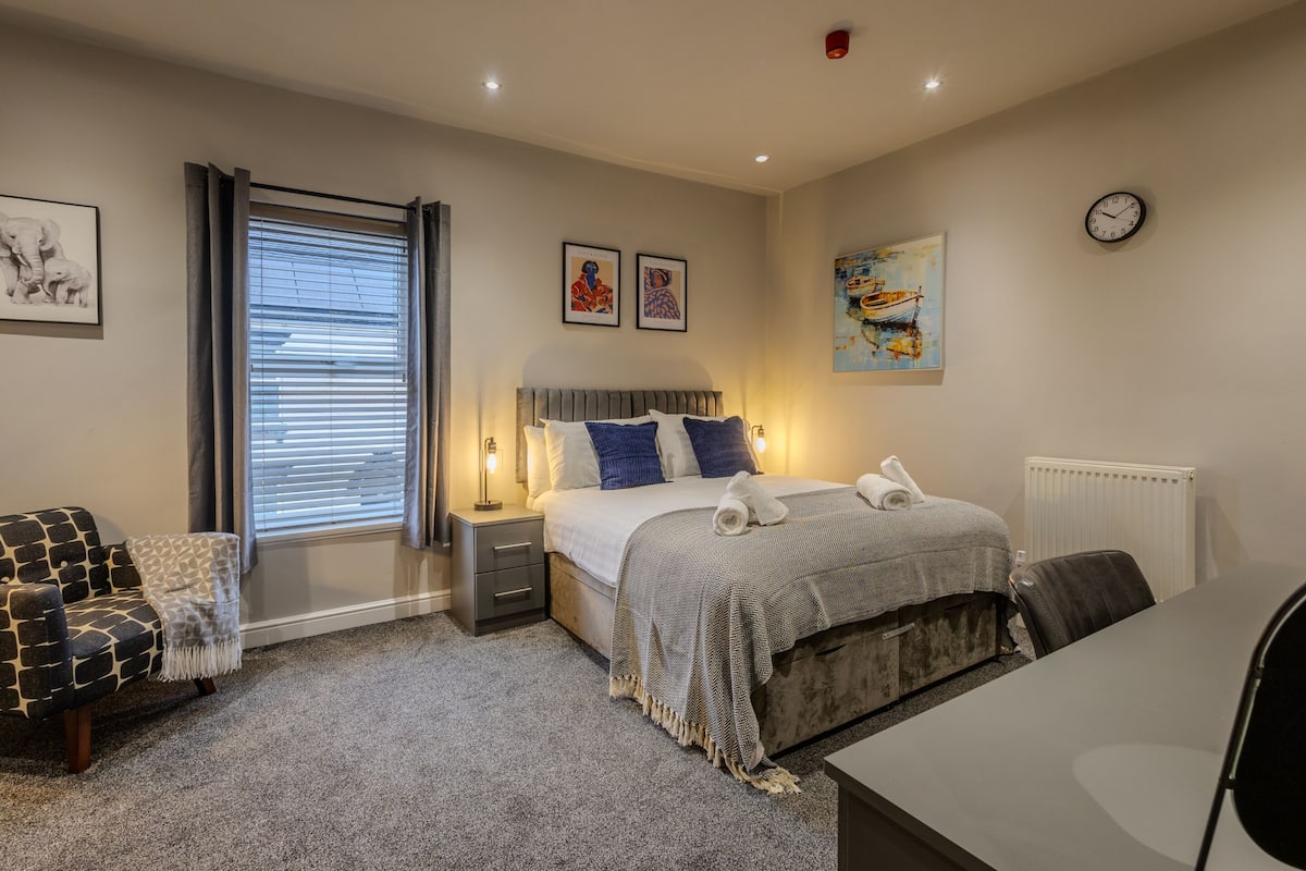 A bedroom with a bed, bedside tables, a chair, a desk, a radiator, and artwork on the walls.
