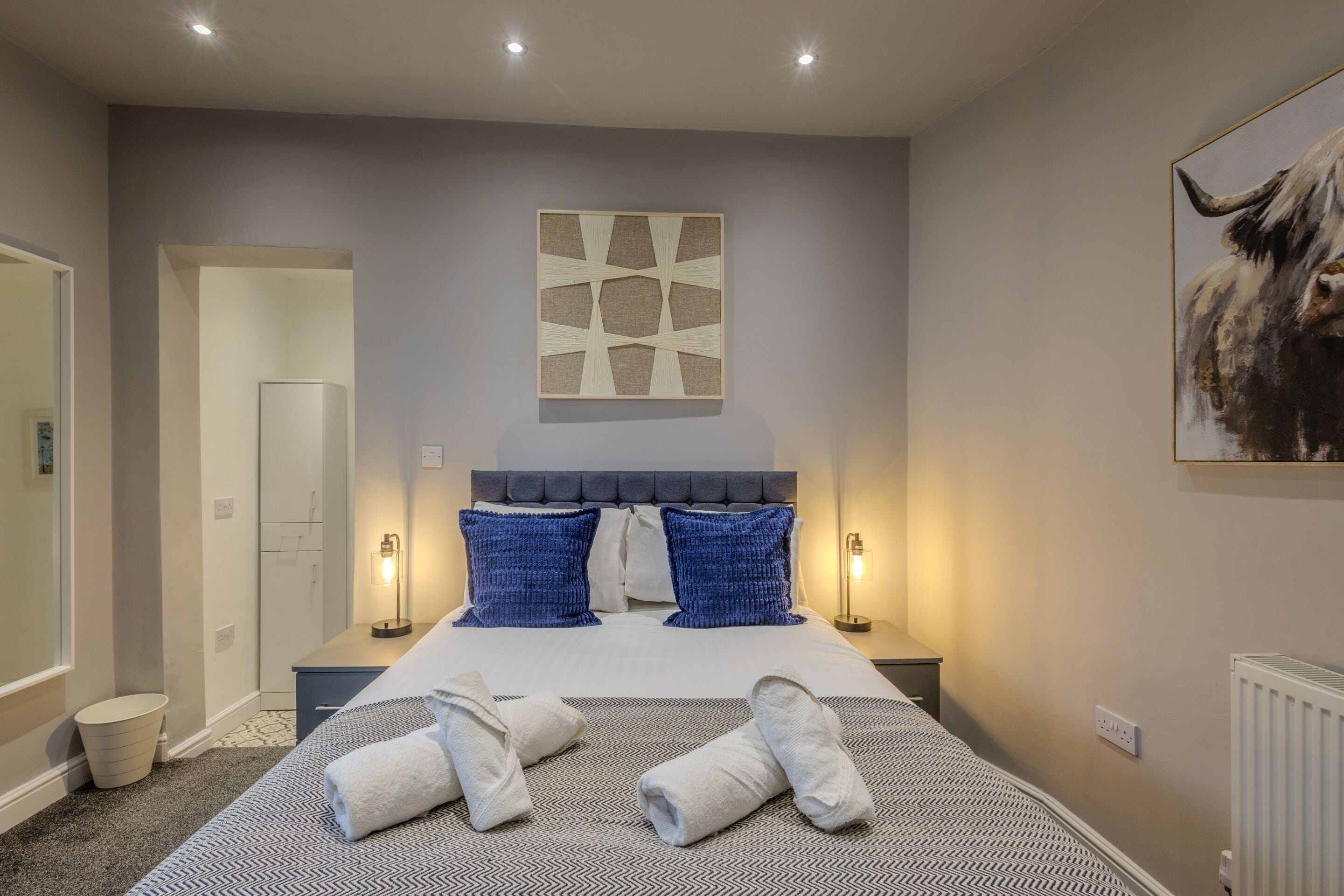 A hotel room with a bed, two bedside lamps, a headboard, a painting, and a radiator.